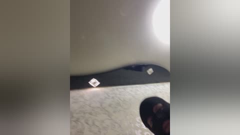 A video of a person with a black headscarf, standing in a carpeted room with a white wall and a black-framed window with diamond-shaped glass. The image is slightly blurred, giving a soft, dreamy effect.