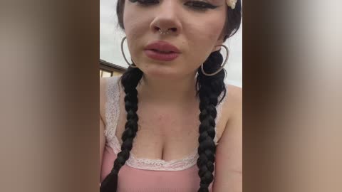 Video of a young woman with pale skin, dark braided hair, pink top, silver hoop earrings, and a septum ring. She's indoors with a blurred background.