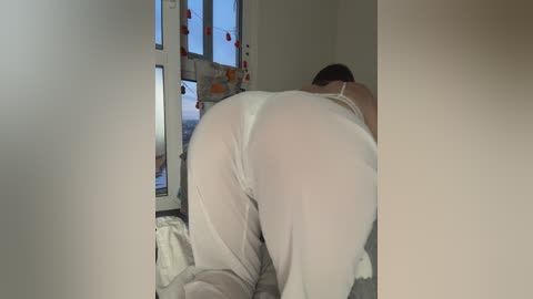 Video of a woman in white lingerie kneeling on a bed, showing her large buttocks through sheer fabric, in a dimly lit room with a window displaying street lights outside.