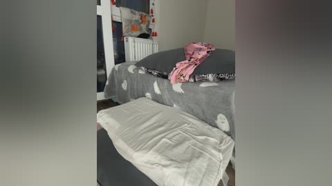 Video of a modest bedroom with a gray bedspread, white pillow, pink pajamas, and a stuffed animal on a white ottoman.