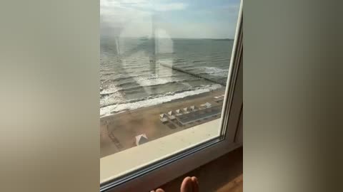 Video of a beach scene viewed through a partially open window. Sunlight filters through the glass, casting a warm glow on the sandy shore. White beach umbrellas line the horizon, and gentle waves roll in the background.