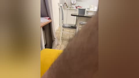 Video of a person's bare arm and torso in a modern kitchen with light wood floors, white cabinets, and a yellow chair in the foreground.