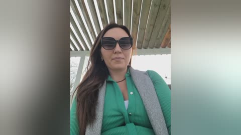 Video of a middle-aged woman with long dark hair, wearing a green cardigan over a white top, black sunglasses, and standing under a wooden pergola.
