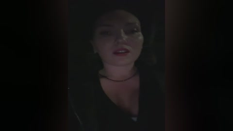 A dimly lit video of a woman with a fair complexion and dark hair, wearing a black top, standing in front of a dark, blurry background.