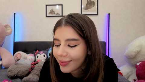 A video of a young Latina woman with long brown hair, wearing black, smiling softly while surrounded by plush toys in a cozy, dimly lit bedroom.