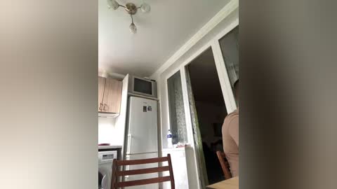 Video of a modern, minimalist kitchen with white cabinets, a stainless steel fridge, wooden chairs, and a beige armchair in a doorway leading to a dimly lit living room.