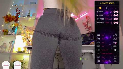 Video of a woman in a gray sports bra and leggings standing in a colorful, well-lit room with shelves filled with flowers and plants.