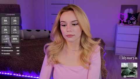 A video of a young, fair-skinned blonde woman in a pastel pink top, seated in a cozy bedroom with plush toys and purple lighting.