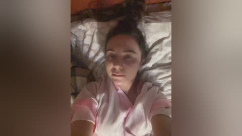 Video of a young woman lying on a bed with messy sheets, wearing a pink shirt, with dark hair, and holding a selfie stick.