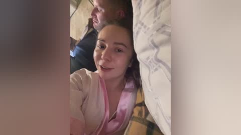 Video of a young woman with light skin, brown hair, and a white robe, peeking through a gap in a white wall, surrounded by a man's face and a beige brick wall.