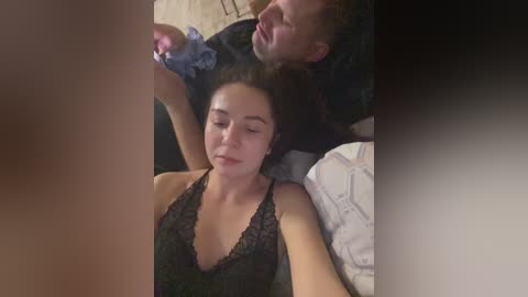 Video of a young woman with light skin, dark hair, and a black lace bra, lying on a bed, with a man holding a pink vibrator beside her.