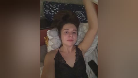 A video of a young woman with light skin, lying on a bed in a dimly lit room. She has brown hair and is wearing a black lace camisole.