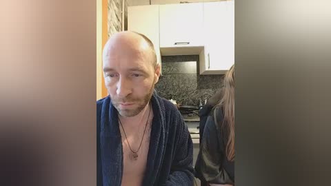 Video of a bald, bearded man in a blue robe, standing in a modern kitchen with white cabinets and dark granite countertops.