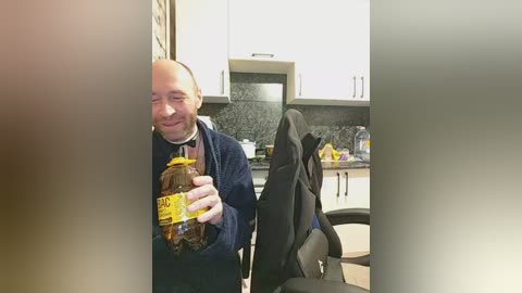 Video of a bald man in a dark jacket, holding a large bottle of dark liquid. He's standing in a modern kitchen with black countertops, white cabinets, and a black chair in the background.