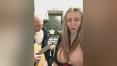 Video of a woman with long brown hair and large breasts, wearing a low-cut top, playing an acoustic guitar in a kitchen with a bald man in a dark shirt.