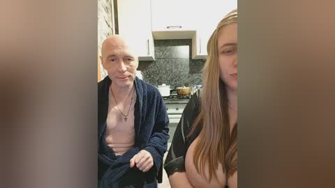 A video shows an elderly man with a shaved head in a robe, standing beside a young woman with long hair, both in a modern kitchen.