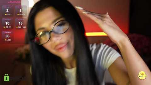 Video of a smiling woman with long black hair, wearing glasses, adjusting her hair. Background shows a dimly lit room with a red light.