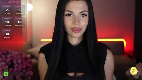 Video of a fair-skinned woman with long black hair, wearing a black sleeveless top, in a dimly-lit room with pink flowers and red lighting.