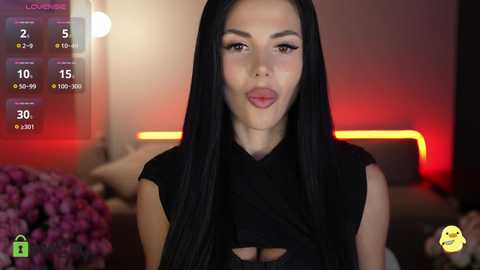 Video of a young woman with long black hair, fair skin, and a slim physique, wearing a black dress. She stands in a dimly lit room with blurred, warm-toned background.