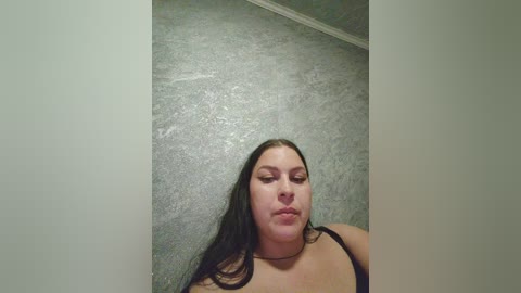 Video of a plus-sized woman with long, straight black hair, fair skin, and a neutral expression, standing against a textured, gray wall. She wears a black tank top, and the image is slightly out of focus.