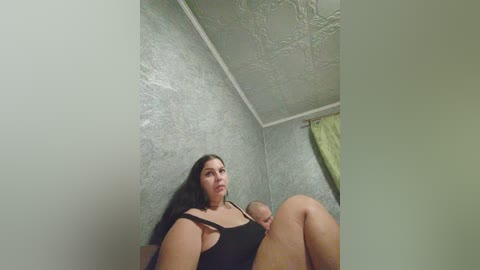 A low-angle video shows a young woman with long dark hair, wearing a black tank top, sitting on a bed with a baby in her arms. The room has grey textured walls and a green curtain.