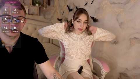 A video of a woman with fair skin, light brown hair, wearing a white lace top and beige skirt, sitting in a pink chair, with a man in a black polo shirt in the background.