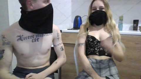 Video of a shirtless man with tattoos and a black mask, seated next to a masked woman with blonde hair, wearing a black crop top and plaid skirt, in a kitchen with white tiles and a blue kettle.