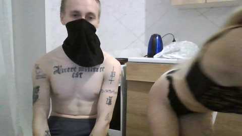 Video of a shirtless, tattooed man wearing a black face mask, sitting on a bathroom counter with a mirror reflection of a woman in black lingerie.