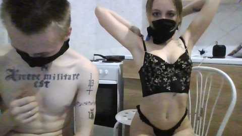 Video of a young woman with a mask and black lingerie, posing in a kitchen with a shirtless man.