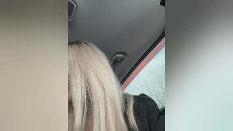 Video of a person with long, straight blonde hair partially obscuring their face, wearing a black jacket, taken from inside a car with a pink and white interior.