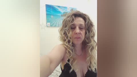 Video of a middle-aged woman with long, wavy, blonde hair, wearing a black lace bra. She is indoors, partially obscured by a white wall, with a blue beach scene painting visible in the background.