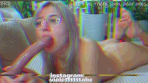 A digital image of a nude, fair-skinned woman with glasses performing oral sex on a large, erect penis. Background features a wicker couch and colorful objects, with a \"Instagram: violer_titties\" watermark.