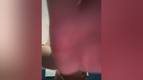 Video of a blurred pink object partially obscuring the face of a person with light skin, standing in a dimly lit room with a white wall and blue floor.