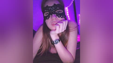 Video of a fair-skinned woman with long brown hair, wearing a black lace masquerade mask, a black wristwatch, and a strapless top, in a purple-lit room, with a seductive, intimate pose.