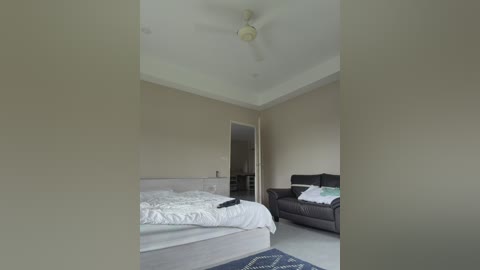 Video of a minimalist bedroom with beige walls, a white bed, a dark gray couch, and a small TV. The room is well-lit, featuring a ceiling fan.