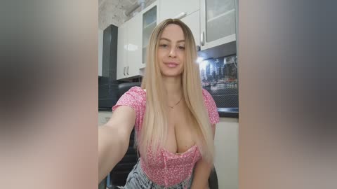 Video of a blonde woman with fair skin, wearing a low-cut pink top revealing cleavage, in a modern kitchen with white cabinets and a dishwasher.