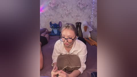 A video of a fair-skinned woman with glasses, lying on a bed, wearing a white shirt, holding a pillow, surrounded by plush toys and fairy lights in a dimly lit room.