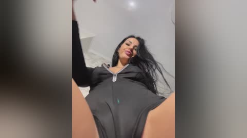 Video of a Latina woman with long black hair, wearing a black bodysuit, standing in a dimly lit room with white walls. She is looking down at the camera, legs spread apart, emphasizing her curvy figure.