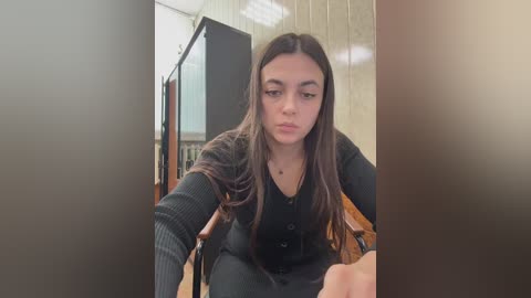 Video of a young woman with long dark hair, wearing a black sweater and jeans, sitting in a dimly lit room with wooden paneling and a television.