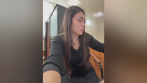 Video of a young woman with long, dark hair, wearing a black sweater, sitting on a wooden bench in a dimly lit hallway with beige walls.