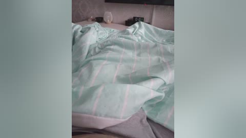 A video shows a bed with a light green and pink striped duvet, partially pulled back to reveal a gray pillow. The background features a beige wall and a dark flat-screen TV mounted above.
