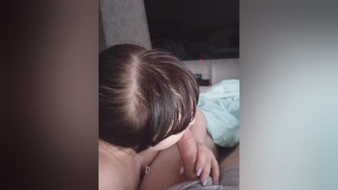 A video of a woman with brown hair and a light skin tone performing oral sex on a man, captured in a dimly-lit, indoor setting with a beige couch and bed in the background.