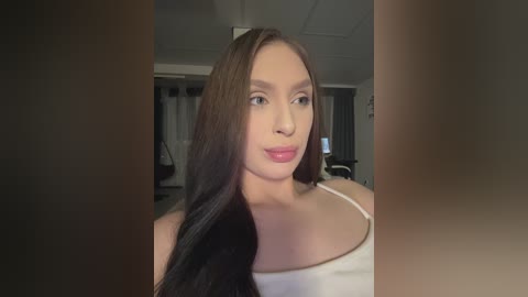 Video of a fair-skinned woman with long, straight, dark brown hair, wearing a white tank top. She has a slender build and a neutral expression. The background is a dimly lit, modern room with white walls and a TV visible.