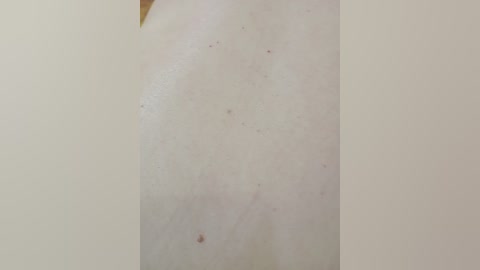 A close-up video of a pale, smooth, slightly bumpy skin surface, possibly on a person's back or side, with small red spots and light freckles. The background is a plain, off-white wall.