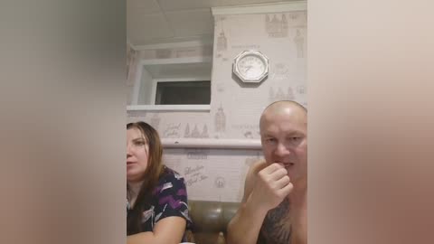 Video of a bald, muscular man with tattoos, shirtless, and a woman with brown hair, wearing a purple-patterned blouse, sitting in a room with wallpaper and a clock on the wall.
