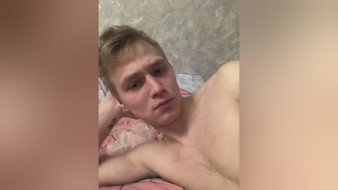 Video of a shirtless, light-skinned, fair-haired young man with a lean physique, resting on a bed with a patterned pink and blue sheet. Background shows textured, gray wall. He has a pensive expression, head tilted slightly to the side.