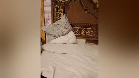 A video of a neatly made bed with white sheets and a patterned pillow, set against a richly decorated, ornate wooden headboard. The background includes a hint of a patterned wall.