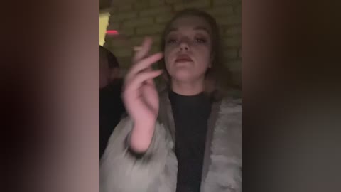 A video of a woman with fair skin, brown hair, and light makeup, dressed in a black top and beige jacket, gestures with her left hand while standing in a dimly lit brick-walled room.