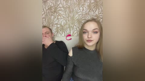 Video of a smiling blonde woman in a gray top and a man with short hair in a black shirt, both standing in a room with a wall mural of palm trees.