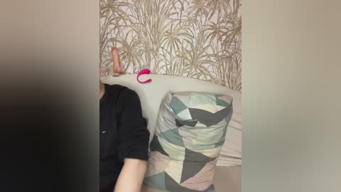 A video captures a person lying on a bed, with their head covered by a white blanket, against a wallpaper featuring a tropical leaf pattern. The person is wearing a black shirt and green, geometric-patterned bedding.